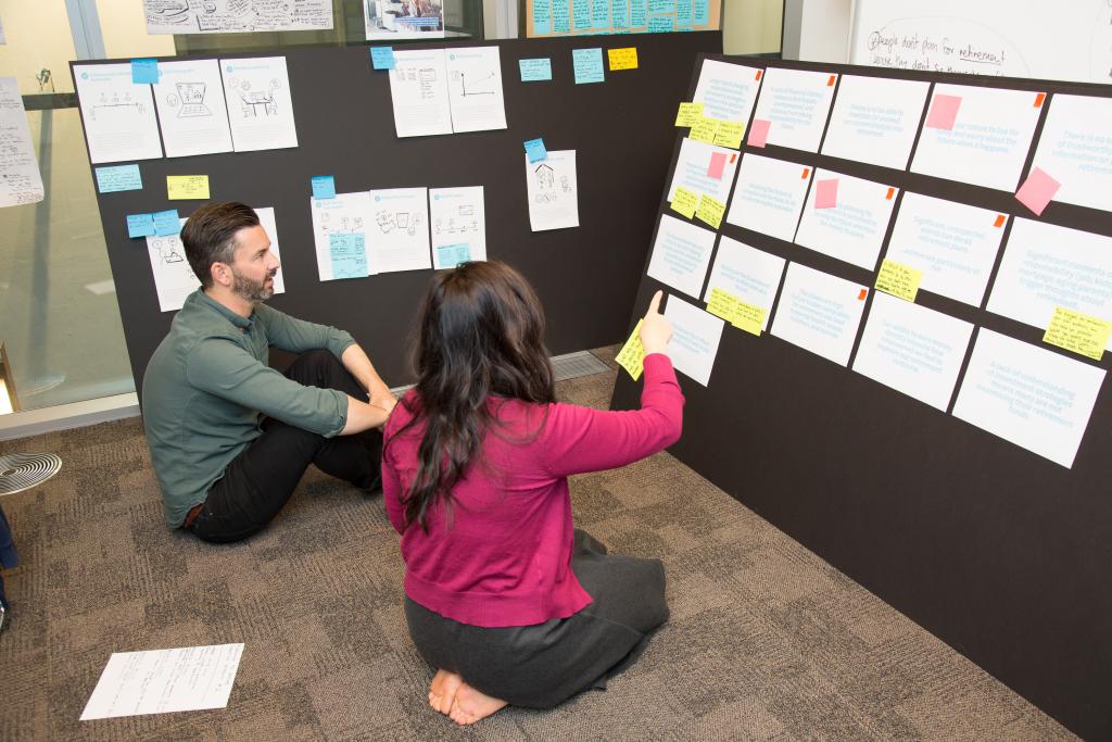 Two people sitting in front of two cardboard walls with a lot of print-outs and sketches, stuck on them.