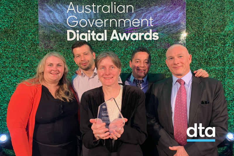 5 people in front of a plant wall. One is holding an award.