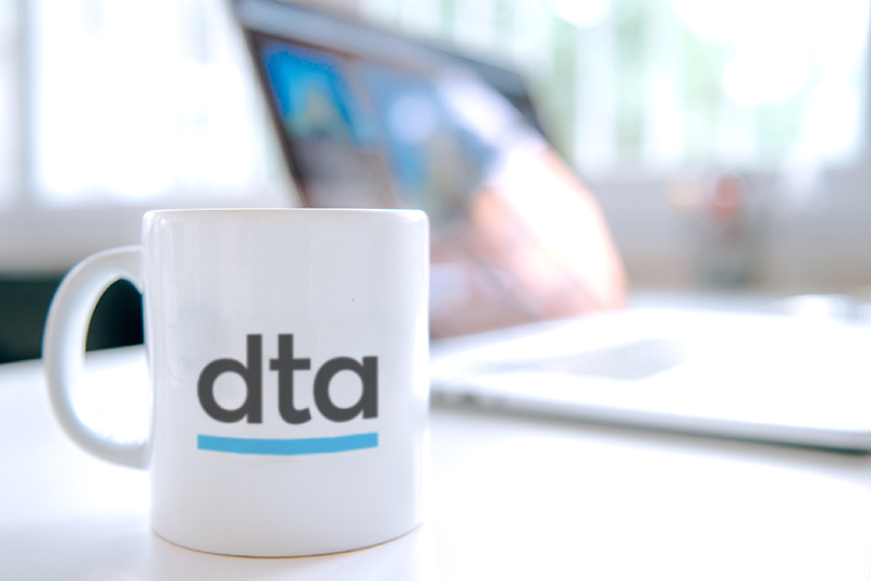 A white mug with 'DTA' written on it. In the background is a laptop.