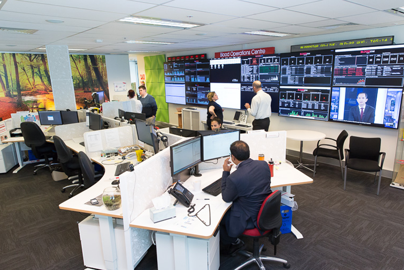 Office desks in front of a wall of digital screens showing various data.