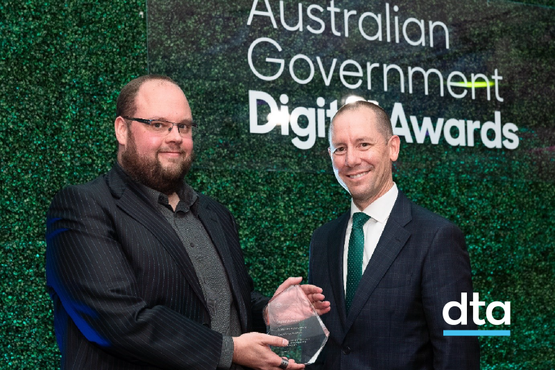Two men standing in front of a plat wall. One is receiving an award from the other.