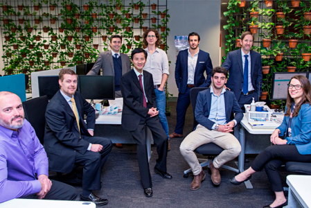 A group of men and women in business attire standing and sitting in front of a wall covered in flowering plants.