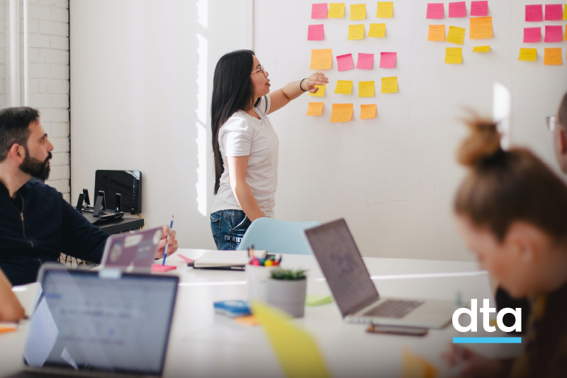 Woman in an office with co-workers, pointing at post it notes on a wall.